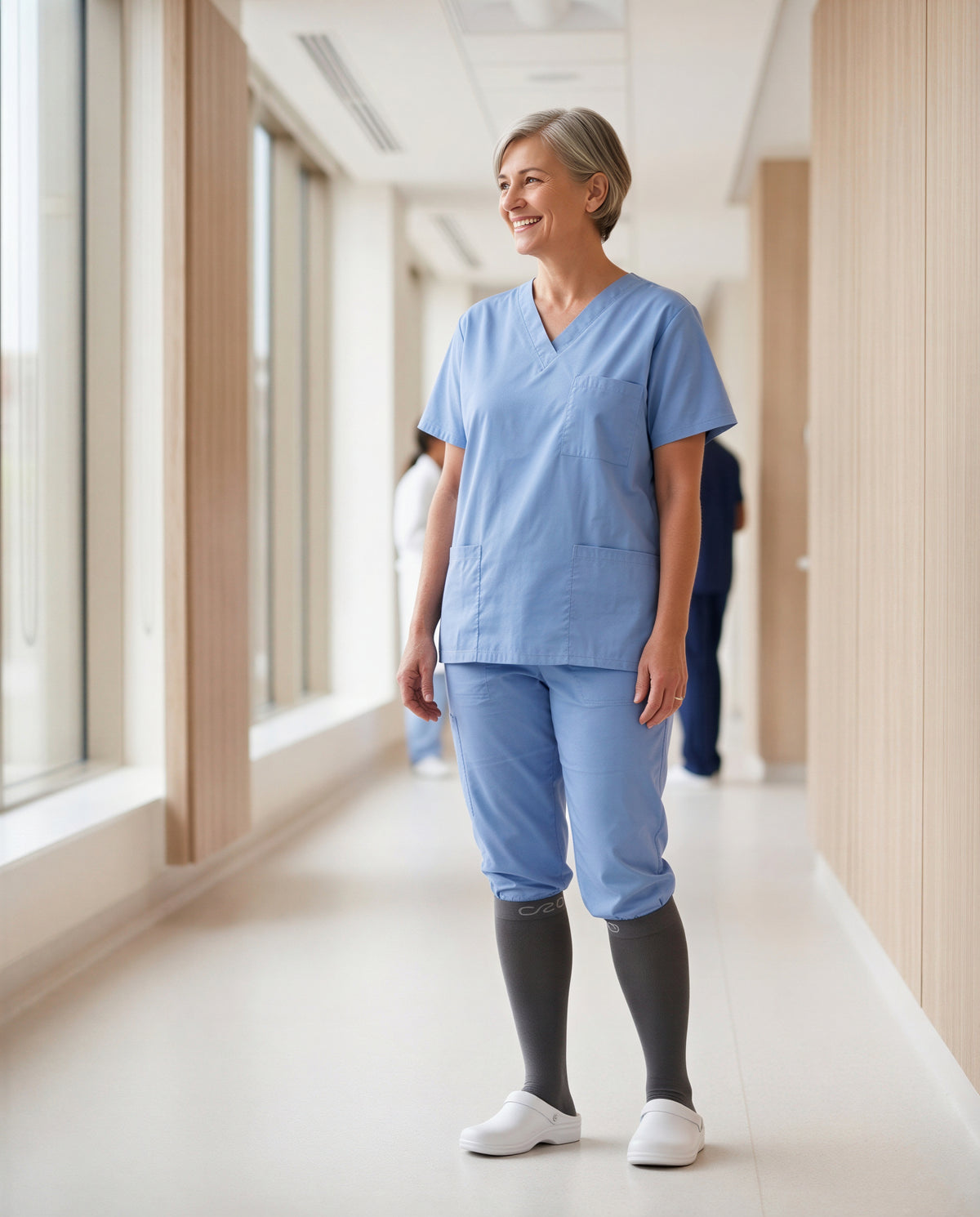 Smiling nurse standing in a bright hospital corridor in blue scrubs and grey knee-high compression socks
